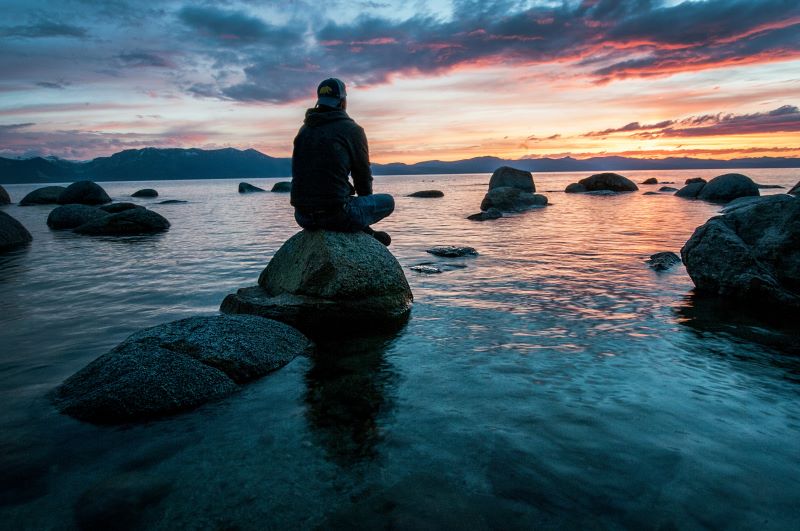 a man sits peacefully on a large rock surrounded by water while looking at the sunset